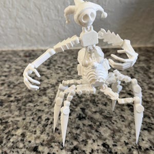 May include: A white, articulated figurine resembling a jester or court jester. The figure has a pointed hat, a skeletal face, and jointed limbs. It has multiple legs and arms, and is posed as if it is dancing or moving.