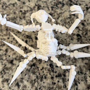 Puede incluir: Una figura blanca articulada que se asemeja a una ara&ntilde;a, con una cara sonriente y extremidades articuladas. La figura tiene m&uacute;ltiples patas puntiagudas y brazos que se pueden posar. El dise&ntilde;o parece estar impreso en 3D, con l&iacute;neas de capa visibles.
