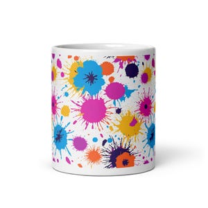 Puede incluir: Taza de cerámica blanca con un diseño de salpicaduras de pintura de colores. El diseño presenta salpicaduras de pintura azul, rosa, amarillo, naranja y morado.