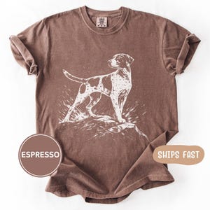 May include: Espresso-colored t-shirt featuring a white graphic of a dog standing on a rock. The dog has brown spots. The shirt has a crew neck and short sleeves. The shirt is made of a soft, comfortable material.