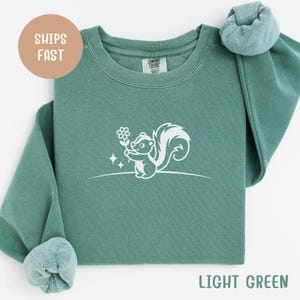May include: Light green sweatshirt featuring a white graphic of a skunk holding a flower. The skunk design is on the front of the sweatshirt. The text "LIGHT GREEN" is at the bottom of the image. The sweatshirt has a crew neck and long sleeves.