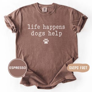 以下が含まれることがあります： 「life happens dogs help」の白文字と足跡がプリントされたエスプレッソカラーのTシャツ。クルーネックのシャツで、袖はロールアップされています。Comfort ColorsブランドのTシャツです。