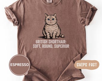 May include: An espresso-colored short-sleeve t-shirt featuring a graphic of a seated British Shorthair cat with blue eyes. Below the cat, the text reads: BRITISH SHORTHAIR: SOFT, ROUND, SUPERIOR. The shirt has a crew neckline and a relaxed fit.