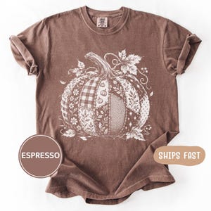 Puede incluir: Una camiseta de manga corta color marrón espresso con un gran gráfico blanco de una calabaza. El diseño de la calabaza está compuesto por varios patrones de patchwork, incluyendo cuadros, flores, espiga y lunares, con hojas arriba.