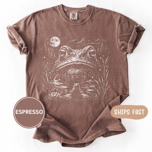 Peut inclure: T-shirt couleur expresso avec une illustration blanche d'une grenouille dans un étang sous la pleine lune. Le motif comprend des nénuphars et des herbes hautes. Le t-shirt a un col rond et des manches courtes.