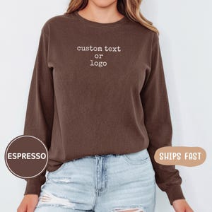 May include: A dark brown long-sleeve crewneck sweatshirt featuring the white text custom text or logo printed on the centre chest. The sweatshirt has a ribbed neckline and cuffs, paired with light blue distressed denim jeans with visible rips.