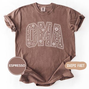 Puede incluir: Camiseta color espresso con la palabra "OMA" en letras blancas llenas de flores. La camiseta tiene cuello redondo y mangas cortas. La palabra "Espresso" está en un círculo marrón en la parte inferior izquierda.
