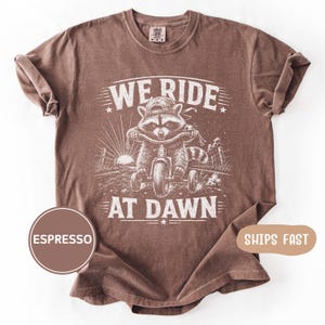 May include: An espresso-colored short-sleeve t-shirt featuring a white graphic print of a raccoon wearing a baseball cap and riding a tricycle. The text WE RIDE AT DAWN is printed in white above and below the raccoon illustration.