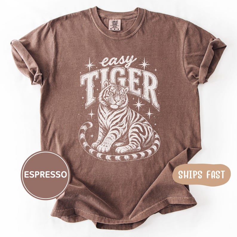 May include: Espresso-colored t-shirt featuring a seated tiger illustration with the text "easy TIGER" in a distressed white font. The design includes star accents. The shirt has a crew neck and short sleeves, with rolled-up cuffs.