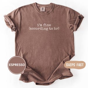 May include: A short-sleeved espresso brown t-shirt with a crew neckline. The center of the chest features white text that reads i'm fine (according to hr) in a simple, lowercase font. The garment has a relaxed fit with rolled sleeves.