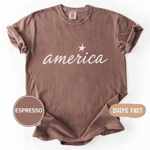 May include: Espresso-colored t-shirt with the word "america" in white script and a white star above the word. The shirt has a crew neck and short sleeves. The shirt is made of a soft, comfortable material.