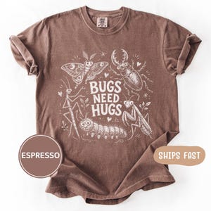 May include: An espresso-colored short-sleeve t-shirt featuring a white graphic print of various insects including a moth, beetle, caterpillar, and praying mantis. The center text reads BUGS NEED HUGS, surrounded by small heart and star accents.