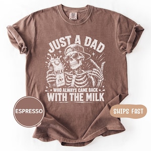 May include: Espresso-colored t-shirt with a graphic of a skeleton wearing sunglasses and a baseball cap, holding a milk carton. The text reads "JUST A DAD WHO ALWAYS CAME BACK WITH THE MILK."