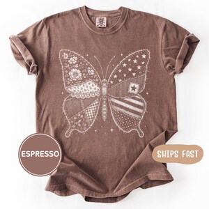 Könnte beinhalten: Espressofarbenes T-Shirt mit detailliertem Schmetterlingsdesign. Die Schmetterlingsflügel bestehen aus verschiedenen Mustern, darunter Blumen, Sterne, Streifen und Patchwork. Das T-Shirt hat einen Rundhalsausschnitt und kurze Ärmel.