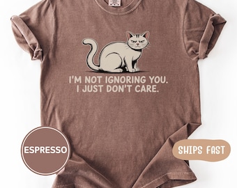 May include: A brown short-sleeved t-shirt featuring a graphic of a sitting cat with a grumpy expression. Below the cat, the text reads: I'M NOT IGNORING YOU. I JUST DON'T CARE. The shirt has a crew neckline and a relaxed fit, with a label at the neck.