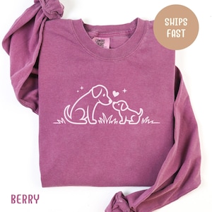 May include: Berry-coloured long-sleeve shirt featuring a white graphic of two dogs, an adult and a puppy, touching noses. The design includes grass, stars, and a heart. The shirt has a crew neck and the text "Comfort Colors" on the tag.