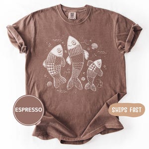 Puede incluir: Una camiseta marrón de manga corta con un gráfico de líneas blancas de tres peces con ojos de botón y patrones de estilo patchwork. Los peces están rodeados de pequeñas burbujas y conchas. La camiseta tiene cuello redondo y etiqueta Comfort Colors.