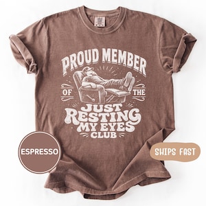 Könnte beinhalten: Espressofarbenes T-Shirt mit einem Aufdruck, der lautet: „PROUD MEMBER OF THE JUST RESTING MY EYES CLUB“. Das Design zeigt eine Person, die sich in einem Stuhl zurücklehnt. Das Shirt hat einen Rundhalsausschnitt und kurze Ärmel.