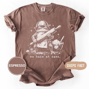 Könnte beinhalten: Espressofarbenes T-Shirt mit einer skurrilen Illustration eines Frosches mit Kochmütze und Schürze, der ein Nudelholz hält. Der Text "we bake at dawn" ist unter dem Frosch aufgedruckt. Das Shirt ist aus weichem Material.