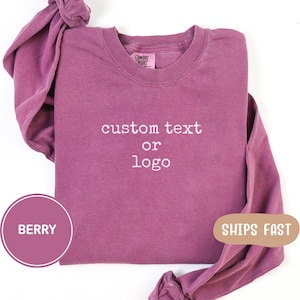 May include: A berry-coloured long-sleeve crewneck sweatshirt with a Comfort Colors label at the inner neckline. The front of the garment features the embroidered white text custom text or logo centred on the chest area in a simple font.