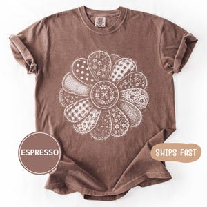 Puede incluir: Camiseta color espresso con un gran diseño floral blanco. Los pétalos de la flor muestran varios patrones, incluyendo lunares, cuadros vichy e estampados florales. La camiseta tiene cuello redondo y mangas enrolladas, ofreciendo un estilo casual.
