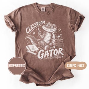 May include: A brown short-sleeved t-shirt featuring a white graphic of an alligator wearing glasses, holding a pencil, and sitting next to a stack of books and a notebook. The text reads CLASSROOM GATOR, LESSONS, IDEAS, STUDENTS, BRIGHTER TOMORROWS.