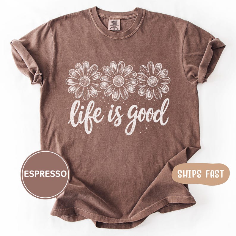 May include: Espresso-colored t-shirt featuring three daisy illustrations above the phrase "life is good" in white script. The shirt has a crew neck and short sleeves. The shirt is made of a soft, comfortable material.