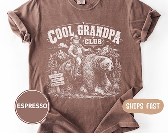Könnte beinhalten: Ein braunes kurzärmeliges T-Shirt mit einem weißen Grafikdruck, der eine Person auf einem Bären reitend zeigt. Der Text lautet COOL GRANDPA CLUB, mit den Wörtern LEGENDARY, RELAXED und UNSTOPPABLE auf einem Schild. Rundhalsausschnitt.