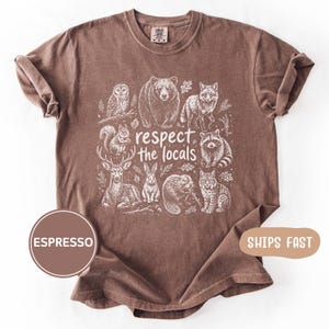 May include: Espresso-colored t-shirt featuring a white graphic of various woodland animals, including a bear, fox, and raccoon, with the text "respect the locals". The shirt has rolled sleeves and a crew neck.