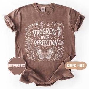 以下が含まれることがあります： エスプレッソカラーの半袖Tシャツ。白のグラフィックプリントには、花や星に囲まれた毛虫、さなぎ、蝶が描かれています。テキストは、手書き風のスタイリッシュなフォントで「PROGRESS OVER PERFECTION」と記されています。