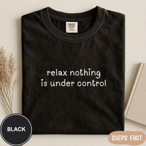 May include: Black t-shirt with the white text "relax nothing is under control". The shirt has a crew neck and a tag that says "Comfort Colors". A black circle with the word "BLACK" is in the lower left corner.