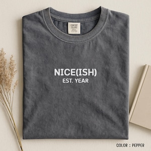 May include: A folded, pepper-coloured t-shirt with the text "NICE(ISH) EST. YEAR" printed in white. The shirt has a crew neck and a tag from Comfort Colors. The shirt is on a neutral background.