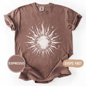May include: A short-sleeve crew neck t-shirt in a solid espresso brown color. The center of the shirt features a large, white, distressed-style graphic illustration of a sun with radiating wavy rays, small stars, and crescent moon accents.