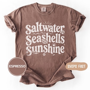 May include: Espresso-colored t-shirt with the words "Saltwater, Seashells, Sunshine" in a white, vintage-style font. The design includes seashell and wave graphics. The shirt has rolled-up sleeves.