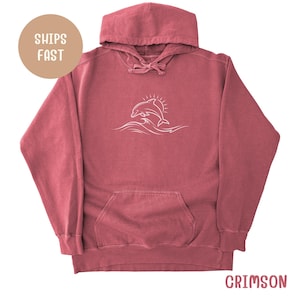 Puede incluir: Sudadera con capucha de color carmesí con bolsillo de canguro y capucha con cordón. Un gráfico blanco de un delfín saltando sobre las olas está centrado en la parte delantera. El texto "SHIPS FAST" está en un círculo en la parte superior izquierda.