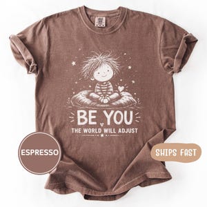 May include: A brown short-sleeve t-shirt featuring a white graphic of a child with messy hair sitting on a cushion. The text reads BE YOU THE WORLD WILL ADJUST. The shirt has a crew neckline and a relaxed fit with rolled sleeves.