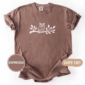 Könnte beinhalten: Ein kurzärmeliges T-Shirt mit Rundhalsausschnitt in einfarbigem Espresso-Braun. Auf der Brust befindet sich eine weiße grafische Illustration einer Eule, die auf einem belaubten Ast sitzt. Das T-Shirt hat eine lockere Passform mit Ärmelaufschlägen.