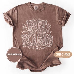 May include: Espresso-colored t-shirt with the words "BE KIND" in a floral design. The text is in a bold, blocky font, with flowers and leaves filling the letters. The shirt has a crew neck and short sleeves.