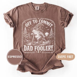 May include: An espresso-coloured short-sleeve t-shirt featuring a white graphic print of a frog wearing a star-patterned wizard hat while riding a scooter. The text reads OFF TO COMMIT DAD FOOLERY, surrounded by a forest landscape and a moon.