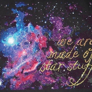 Puede incluir: Bordado a punto de cruz que representa una nebulosa colorida con una inscripción dorada que dice "we are made of star stuff".