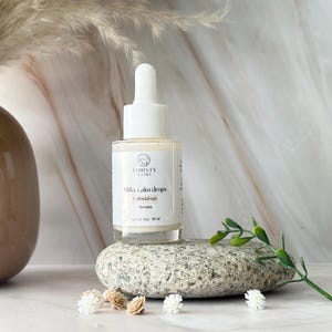 May include: A clear glass bottle of "Milky Calm Drops" serum with a white dropper top. The label features the brand name "THIRSTY KAMEL" and product details. The bottle rests on a gray stone, with small decorative flowers and greenery nearby. The serum is 1 fl oz / 30 ml.