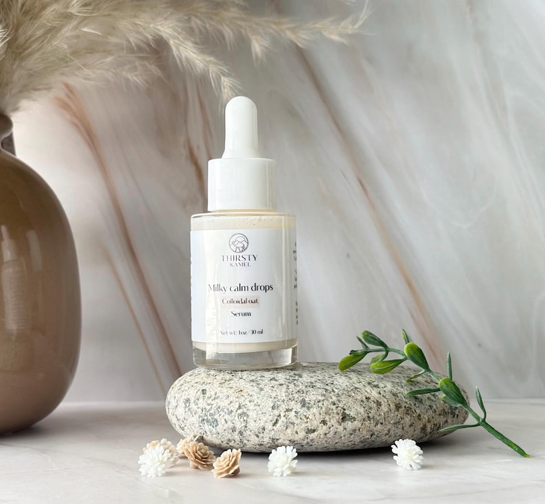 May include: A clear glass bottle of "Milky Calm Drops" serum with a white dropper top. The label features the brand name "THIRSTY KAMEL" and product details. The bottle rests on a gray stone, with small decorative flowers and greenery nearby. The serum is 1 fl oz / 30 ml.