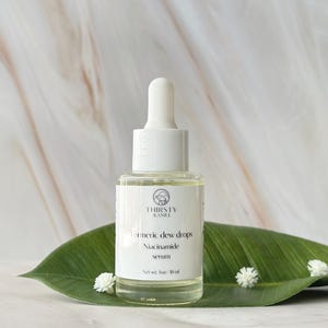 May include: A clear glass bottle of "Turmeric Dew Drops Niacinamide serum" with a white dropper, set on a large green leaf. The bottle has a white label with the brand name "THIRSTY KAMEL" and a logo. The background is a marble pattern.