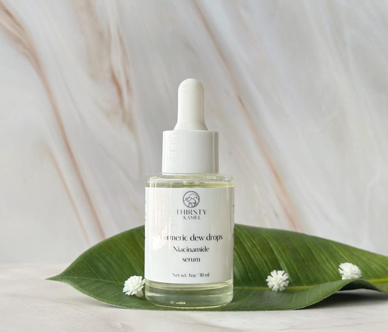 May include: A clear glass bottle of "Turmeric Dew Drops Niacinamide serum" with a white dropper, set on a large green leaf. The bottle has a white label with the brand name "THIRSTY KAMEL" and a logo. The background is a marble pattern.