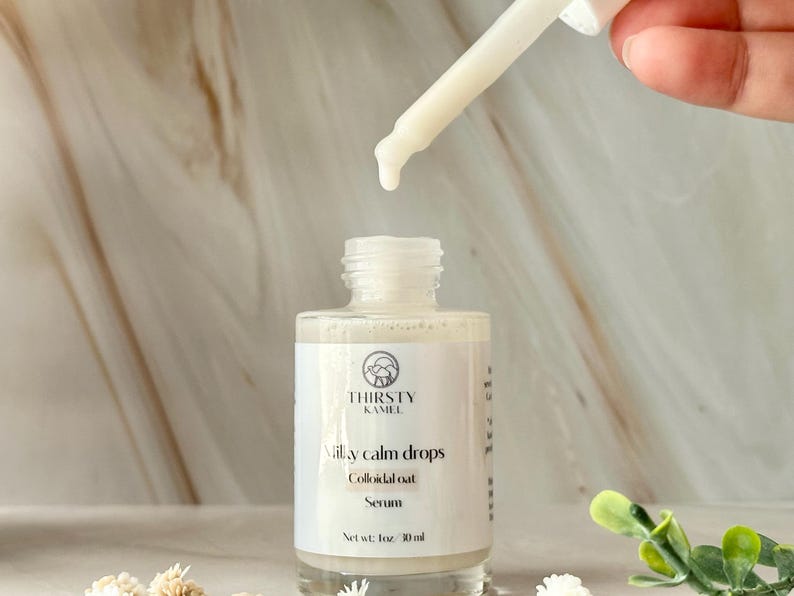 May include: A close-up of a white serum bottle with a dropper dispensing liquid. The bottle is labeled "Milky Calm Drops" and "Colloidal Oat Serum" by Thirsty Kamel. The bottle contains 1 fl oz (30 ml) of serum. The background is a marble-like surface.