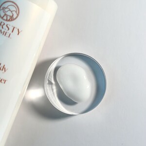 Pu&ograve; includere: Un piccolo ciuffo di crema viso bianca in una ciotola di vetro trasparente. La crema &egrave; liscia e densa, e la ciotola &egrave; rotonda e poco profonda.
