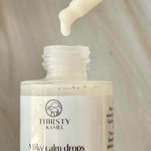 May include: A close-up of a white bottle of "Milky Calm Drops" with a dropper dispensing a milky liquid. The bottle has a label with the text "Thirsty Kamel" and a camel logo. The background is a light, marble-like surface.