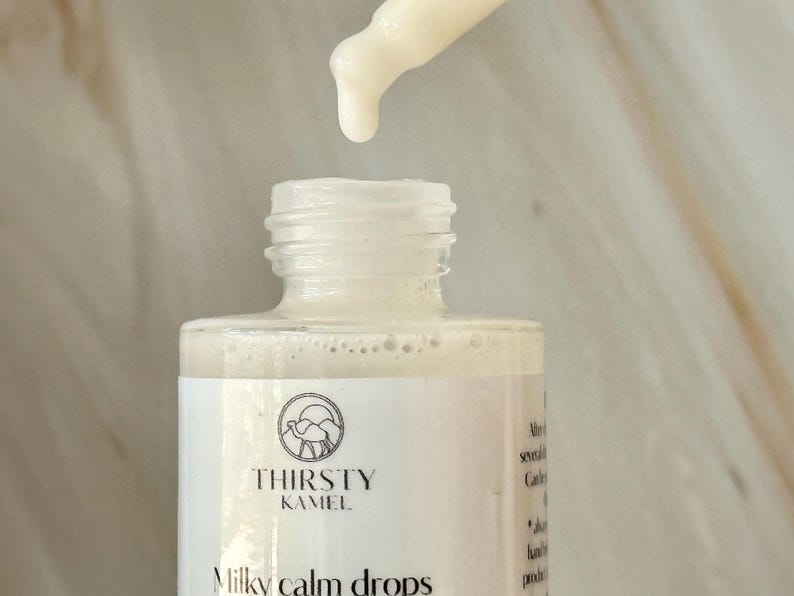 May include: A close-up of a white bottle of "Milky Calm Drops" with a dropper dispensing a milky liquid. The bottle has a label with the text "Thirsty Kamel" and a camel logo. The background is a light, marble-like surface.