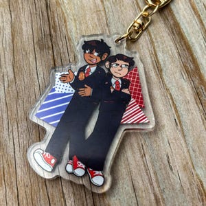 Puede incluir: Un llavero de acrílico transparente con dos personajes de dibujos animados con traje y zapatos rojos. Los personajes están de pie sobre un fondo de formas geométricas rojas, blancas y azules.