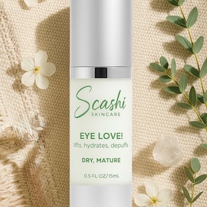 Op de afbeelding: Een flesje huidverzorgingsproduct voor de ogen met een zilveren dop en witte basis, met het label "Scashi SKINCARE EYE LOVE!" voor de droge, rijpere huid. De fles bevat 15 ml. De achtergrond bevat witte bloemen en groen.